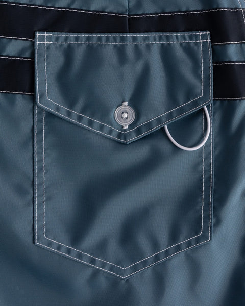 Close-up of a 300 Comp Stripe - Federal Blue/Navy boardshort pocket, featuring white stitching, a flap with round button, and silver side loop—classic details of premium USA-made boardshorts.