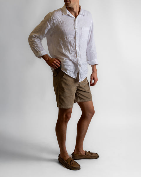 A man stands against a plain background wearing a white long-sleeve button-up shirt, Classic Corduroy Shorts - Acorn with an adjustable drawcord waist, and brown loafers. His right hand is on his hip, face partially out of frame.