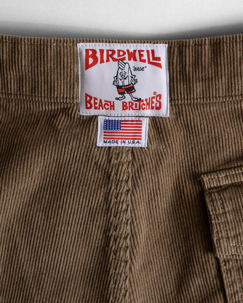 Close-up of Classic Corduroy Shorts - Acorn with two sewn labels: one shows the Birdwell Beach Britches logo and cartoon bird, the other features the American flag, 