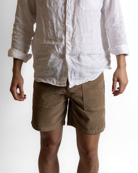 A person wears a white linen button-up shirt with sleeves rolled up and Classic Corduroy Shorts - Acorn, standing against a plain white background. The person's head and lower legs are out of frame.