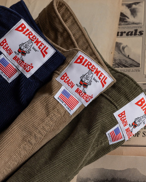 A trio of Classic Corduroy Shorts - Acorn is displayed on vintage papers and newspaper clippings, each pair featuring an adjustable drawcord waist, a signature patch, and an American flag label.