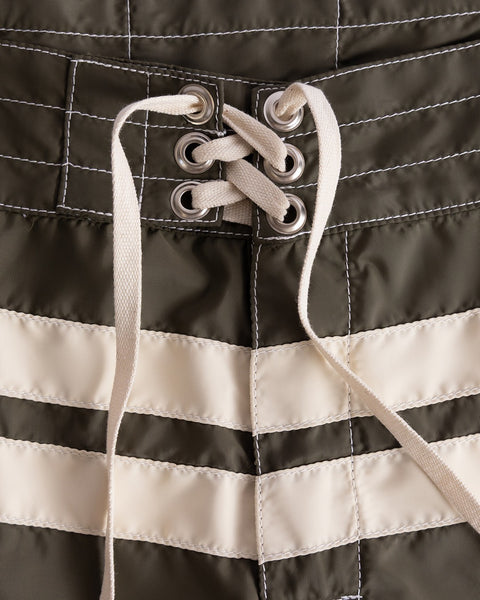 Close-up of 300 Comp Stripe boardshorts in Dark Olive/Bone, featuring white stitched lines, horizontal stripes, and a white lace-up drawstring threaded through metal eyelets at the waist.