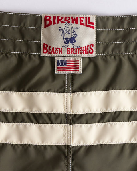 Close-up of the 300 Comp Stripe - Dark Olive/Bone boardshorts, featuring two white stripes, an American flag patch, and the Birdwell Beach Britches label—crafted from SurfNyl and proudly made in the USA.