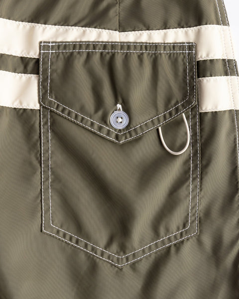 Close-up of the 300 Comp Stripe - Dark Olive/Bone boardshorts pocket with white stitching, flap with button, two beige stripes above, silver D-ring below the flap; proudly made in the USA.