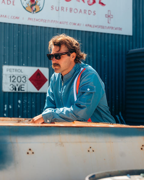 A man with brown hair, a mustache, and sunglasses wears the Racing Jacket 26 in Federal Blue/White/Orange while leaning on a rusty metal surface outdoors near PETROL 1203 3YE and SURFBOARDS signs.