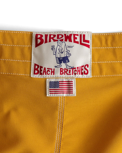A close-up of the 808 Racing Stripes - Gold/Bone, made in the USA, showing the Birdwell Beach Britches label with a cartoon bird and a small stitched American flag below it.