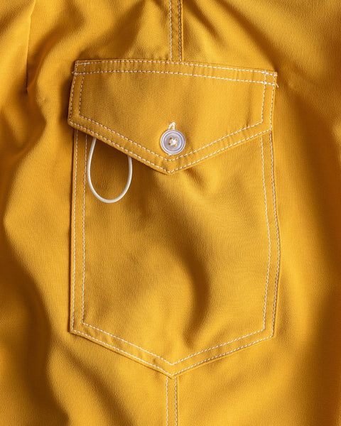 A close-up view of a SurfStretch® fabric pocket on the 808 Racing Stripes - Gold/Bone boardshorts. The yellow pocket features white stitching, a flap with a white button, and a partially visible white loop under the flap.