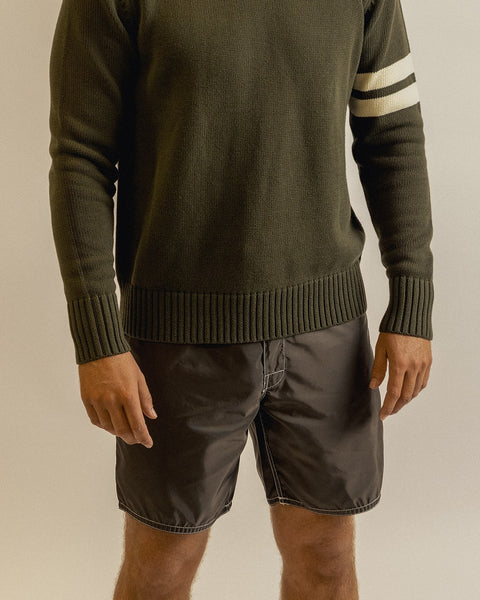 A person wearing a dark green ribbed sweater with two white stripes on one sleeve and brown 310 Boardshorts stands against a plain light background. Their head and lower legs are not visible.