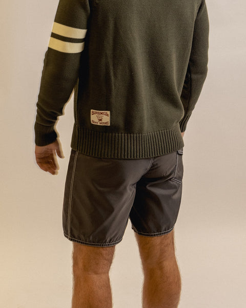A person stands facing away, wearing a dark green sweater with white sleeve stripes and brown 310 Boardshorts. The back patch on the sweater reads BIRKSMILL. The background is neutral and plain.