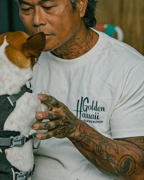 A tattooed man in a Golden Hawaii T-Shirt - Vintage White gently holds a small brown and white dog in a harness, looking at it affectionately.