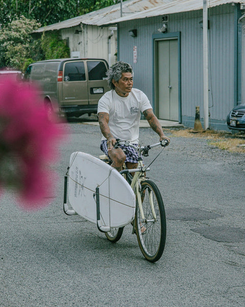 A gray-haired person rides a bike down the street with a white surfboard mounted on the side, wearing a Golden Hawaii T-Shirt - Vintage White, passing a gray building and van as pink flowers blur in the foreground.