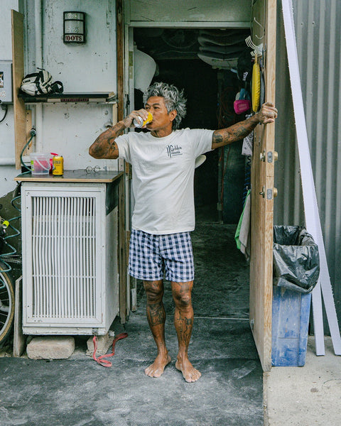 A barefoot, gray-haired man with tattoos stands in a cluttered doorway, wearing the Golden Hawaii T-Shirt in Vintage White and checkered shorts, while drinking from a yellow can surrounded by tools and various objects.