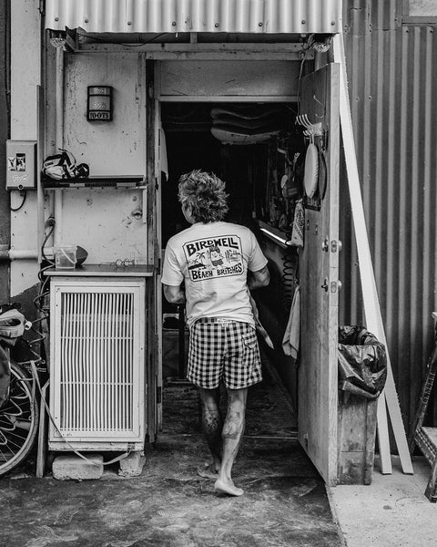 A barefoot person in a Golden Hawaii T-Shirt - Arctic Blue and plaid shorts walks through a cluttered open doorway outdoors, with a bike wheel and trash can nearby. The black-and-white photo captures this candid moment.