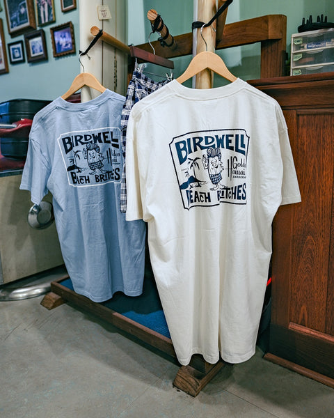 A Golden Hawaii T-Shirt - Arctic Blue with a back graphic hangs on a hanger beside folded shorts in a cozy, vintage-style store.