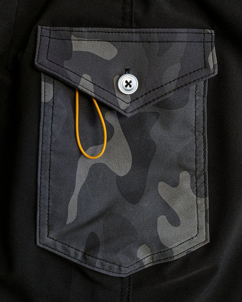 A close-up of a camouflage-patterned pocket with a flap and button on the black 808 Powerbird boardshort. A yellow elastic band is looped and partially tucked under the flap.