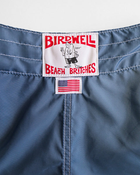 Close-up of the blue waistband on 400 Boardshorts - Federal Blue, featuring the Birdwell Beach Britches logo with cartoon figure and a small quick-dry label displaying the American flag below.