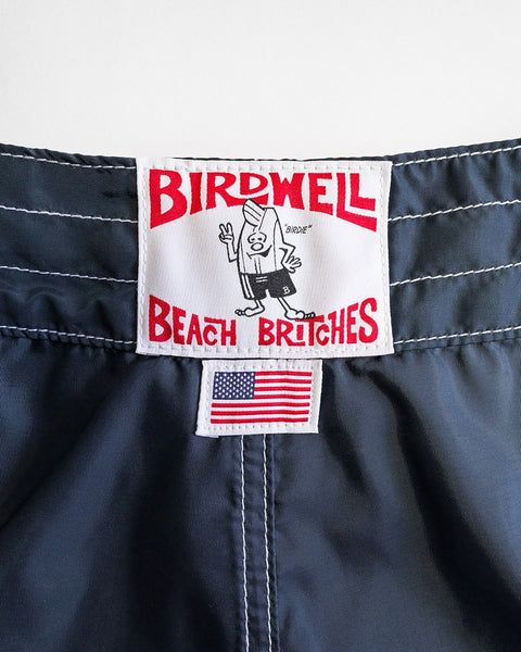 A close-up of the waistband of the 400 Boardshorts - Navy shows a white Birdwell Beach Britches label with a cartoon surfer and an American flag patch, highlighting they’re proudly made in the USA.