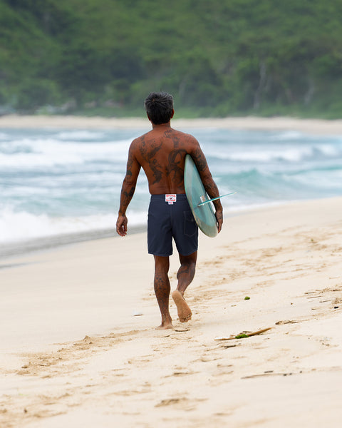 A tattooed man wearing 300 Boardshorts - Navy walks barefoot along a sandy beach, surfboard under his arm, with ocean waves and green foliage in the background.