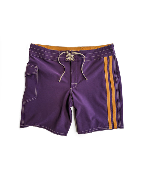 The 808 Racing Stripes - Purple/Gold boardshorts feature a tan waistband, tan drawstrings, two vertical gold stripes on one side, a flap side pocket on the left leg, and visible white stitching throughout.