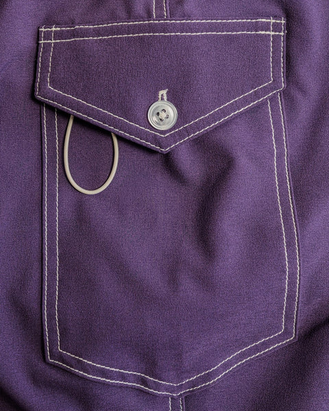 A close-up of the 808 Racing Stripes - Purple/Gold boardshort pocket shows purple SurfStretch fabric with white stitching, a button closure, and a white string loop—premium details found on USA-made boardshorts.