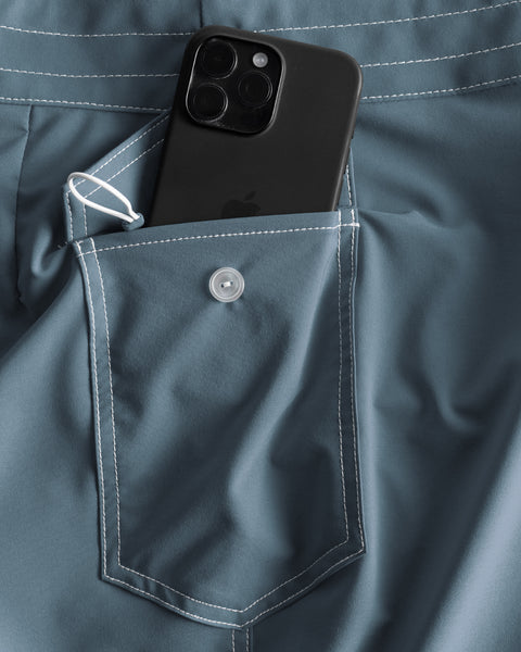 A black smartphone is partially tucked into the buttoned back pocket of the 808 SurfStretch Side Pocket Boardshorts in Federal Blue, featuring white stitching and a small loop beside the pocket.