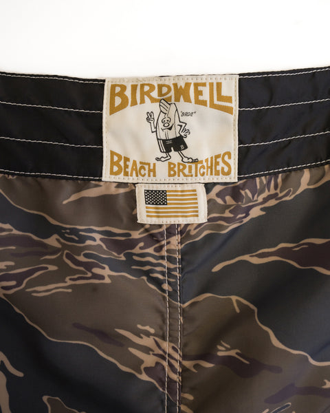 Close-up of the 300 Boardshorts - Tiger Camo with a Birdwell Beach Britches label showing a cartoon character and an American flag tag below, proudly made in the USA.