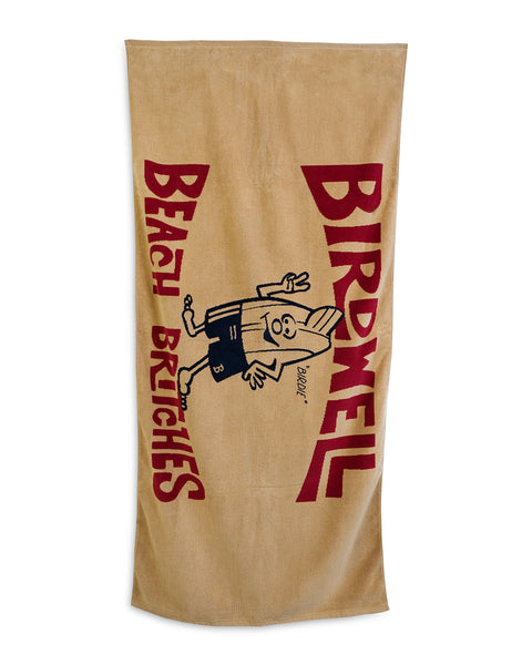 The License Plate Towel - Bone is a large, tan beach towel made in the USA with red BIRDWELL and BEACH BRITCHES text plus a cartoon running surfboard in trunks saying 
