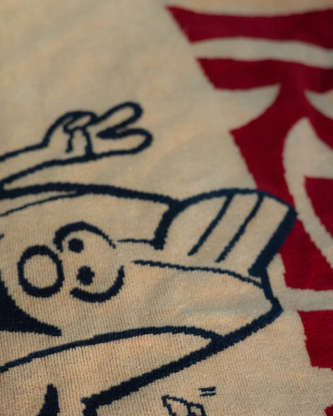 A close-up of the License Plate Towel - Bone, made in the USA, featuring a blue cartoon character flashing a peace sign with part of a large red design in the background.