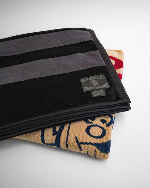 Two folded blankets are stacked, with the top being the Comp Stripe Towel - Black/Gray made from 100% cotton and featuring a visible label, while the bottom is tan with blue and red cartoon-style graphics.