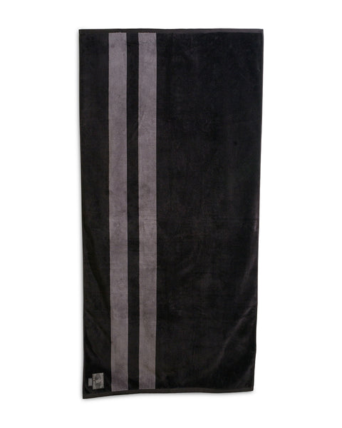 The Comp Stripe Towel - Black/Gray is an oversized 100% cotton towel, featuring three vertical gray stripes on the left and a small sewn patch in the bottom left for added comfort.