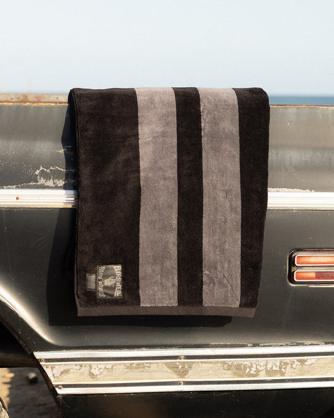 The Comp Stripe Towel - Black/Gray, made from 100% cotton with three vertical gray stripes, hangs over a dark vintage car with the ocean and sky in the background.