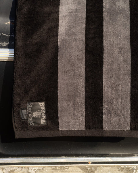 The Comp Stripe Towel - Black/Gray, made from 100% cotton, features two wide gray stripes on a black background. Folded on a dark surface, its corner patch shows the Birdwell logo and American flag, emphasizing its made in USA quality.