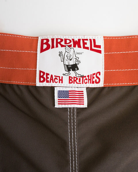 Close-up of the 808 Pop Pocket - Brown/Paprika label with a cartoon character above an American flag tag on brown and orange fabric—classic, water-repellent boardshorts proudly made in the USA.