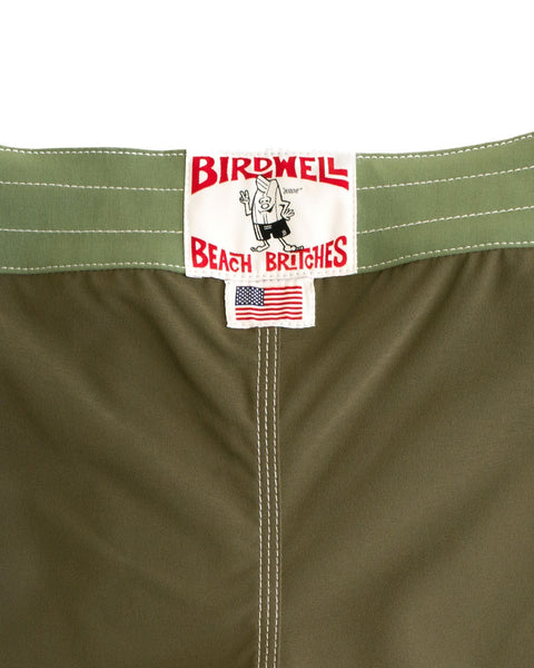 A close-up of the 808 Pop Pocket - Dark Olive/Olive boardshorts shows the Birdwell Beach Britches label and USA flag patch below the waistband, with white stitching accenting their SurfStretch fabric, proudly made in the USA.
