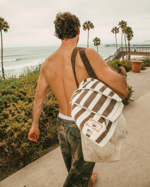 A shirtless man with curly hair, in camouflage pants, walks on a coastal path carrying a striped towel and the Beach Tote - Natural. Palm trees, bushes, and the ocean can be seen in the background.