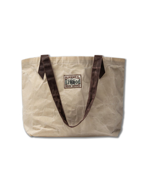 The Beach Tote - Natural is a beige waxed cotton canvas bag with dark brown handles and a rectangular patch. From the Elwood collaboration, its wrinkled design is shown against a white background.