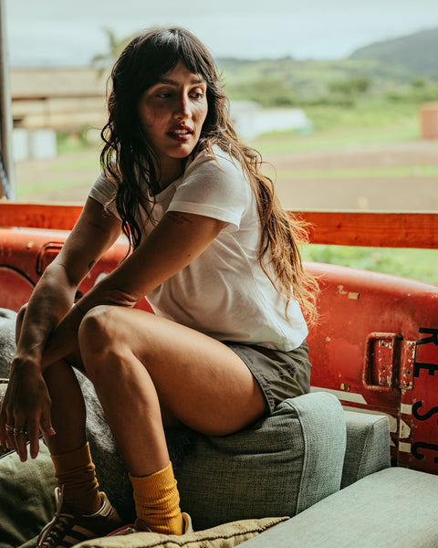 A woman with long wavy hair, wearing a white t-shirt and 400 Boardshorts - Dark Olive, sits on a couch with her knees up, looking to her left. An old red rescue flotation device is behind her, with greenery visible outside.