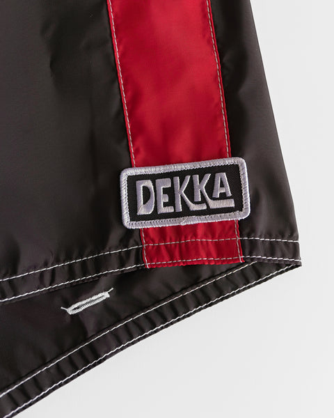 A close-up of the 310 Dekka - Black/Red shows black SurfNyl fabric with a vertical red stripe, a DEKKA patch in white outlined in black and gray, and white stitching along the edge—classic boardshorts style, made in USA.