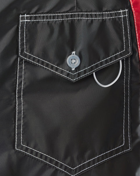 Close-up of the 310 Dekka - Black/Red boardshorts pocket, featuring black fabric, white stitching, a buttoned flap, and a partially visible looped white cord under the flap.