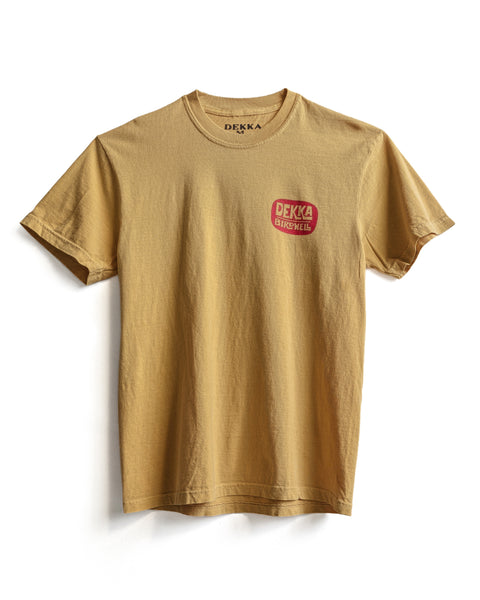 The Birdwell x Dekka T-Shirt - Mustard is a tan, garment-dyed short-sleeve tee made from 100% cotton with a small red DEKKA BICYCLE logo on the upper left chest, shown against a white background.