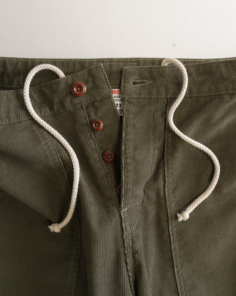 Close-up of the Classic Corduroy Pants - Dark Olive, featuring an olive green corduroy fabric with a white drawstring, brown buttons, and a partially visible tag inside the waistband. The pants are unbuttoned and laid flat against a light background, evoking the relaxed style of Southern California craftsmanship.