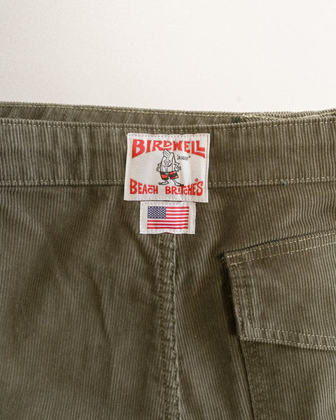 Close-up of the waistband on dark olive Classic Corduroy Pants, made in Southern California, featuring a Birdwell Beach Britches label and a small American flag tag above a back pocket.