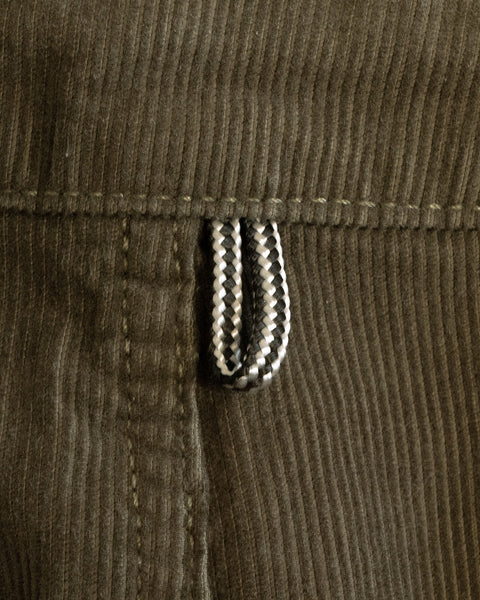 Close-up of a dark olive corduroy fabric, reminiscent of the Classic Corduroy Pants - Dark Olive, featuring a stitched seam and a thick, black-and-white striped cord loop attached. This detail highlights the timeless craftsmanship associated with this iconic style.