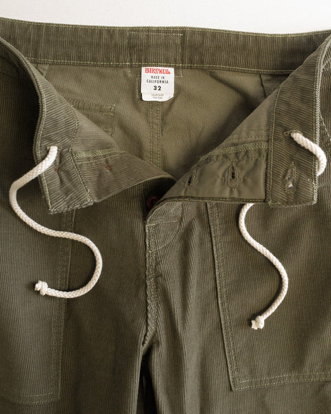 Dark olive Classic Corduroy Pants with a white drawstring, button fly, and visible Brickell brand label on the waistband. Made in Southern California, these corduroy pants are displayed open to reveal the inner waistband and pockets.