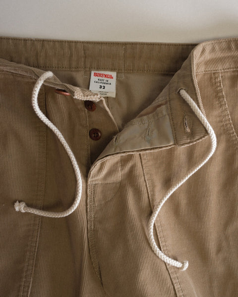 Close-up of the Classic Corduroy Pants - Sand, crafted from 100% cotton corduroy in a tan hue, featuring white drawstrings and brown buttons, with the button fly partially unbuttoned. A label displays “BIRDWELL, California, 32.” The visible waistband and button fly showcase meticulous craftsmanship made in Southern California.