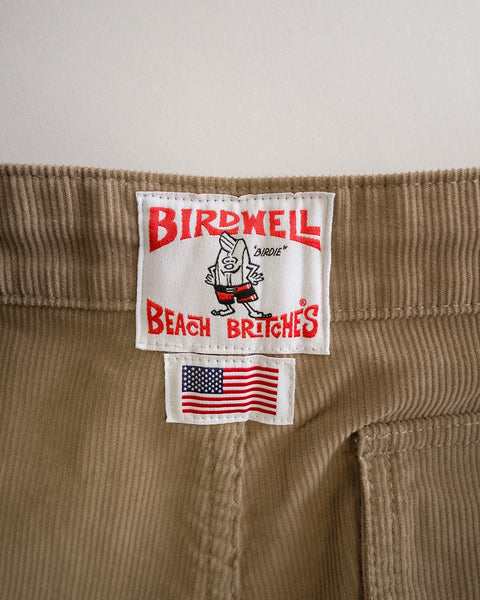 A close-up of the Classic Corduroy Pants - Sand, crafted from 100% cotton corduroy, highlights a Birdwell Beach Britches label featuring a cartoon bird, along with a small tag displaying the American flag beneath it—all proudly made in Southern California.