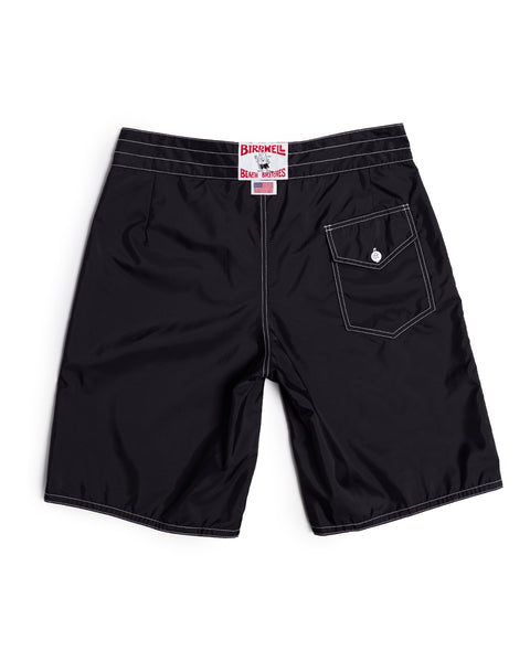 312 Boardshorts - Black: Classic long-length black boardshorts featuring white stitching and a back pocket with a button flap on the right side. A white label with red BIRDWELL text sits above the waistband. These boardshorts are displayed flat, highlighting their iconic SurfNyl construction made in the USA.