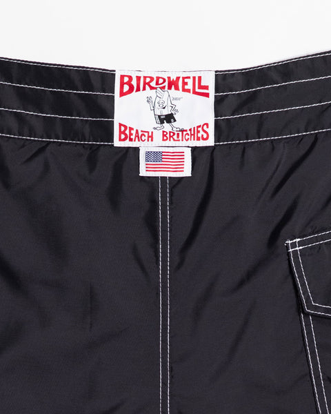 Close-up of black 312 Boardshorts featuring a Birdwell Beach Britches label with a cartoon character, a small American flag tag, and white stitching near a pocket. These long-length 312 Boardshorts are proudly made in the USA.