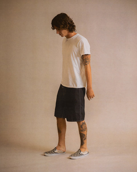 A person with tattoos wears a white t-shirt, black 312 Boardshorts made in the USA, and checkered slip-on shoes, standing against a plain beige background, looking down with arms relaxed by their sides.