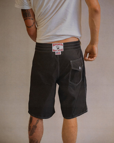 A person stands facing away, wearing a white t-shirt and black 312 Boardshorts with white stitching. A visible red “Birdwell Beach Britches” label is on the shorts, which are made in the USA. Part of a leg tattoo is visible on the left leg.